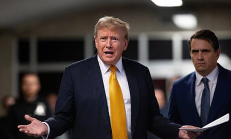 White man wearing black suit and yellow tie gestures with his hands as another man in a suit stands behinds him