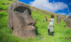 A young man stands on a hillside in front of a colossal statue of a face, taking a picture