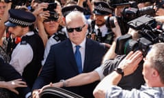 Huw Edwards wearing sunglasses and a suit walks towards a car with an open door surrounded by photographers as police try to clear a way for him