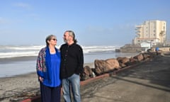 An older white couple gaze at each other, arms around each other, on an asphalt walkway above a beach with a high-rise beyond them.