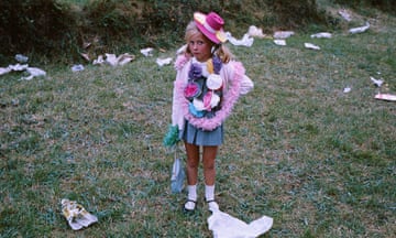 A girl dressed up in a pink hat stands in a field with rubbish behind her