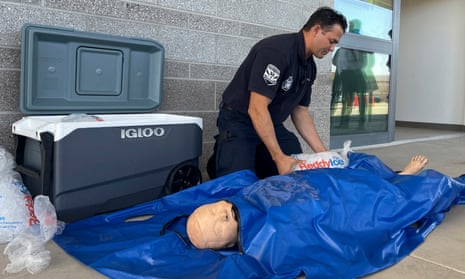 White man in black short-sleeved uniform kneels on sidewalk beside Igloo cooler, holding a bag of ice beside a large blue plastic bag with a peach-colored test dummy inside.