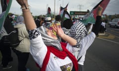 A woman dressed in a black-and-white keffiyeh, white shirt, and red scarf appears to shout and holds a Palestinian flag on a street with a group of other people doing the same.