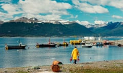 A man in a yellow oilskin jacket on a beach carrying two very huge crabs, while small fishing boats are anchored in an inlet. In the background are snow-topped mountains