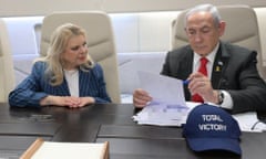 Benjamin Netanyahu sits at a table onboard a jet  looking at some official papers as his wife looks on. In front of Netanyahu is a blue cap with the words 'total victory' on it