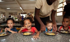 A row of small children eat soup, one helped by an adult