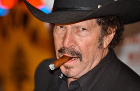 a middle-aged white man with a mustache and chin hair wearing a black cowboy hat smokes a cigar