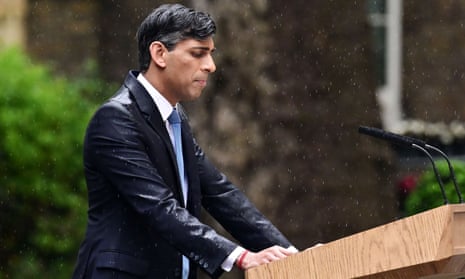 Rishi Sunak from the waist up in a dark blue suit drenched with rain and a light blue tie. He stands behind a lectern as rain pours down.