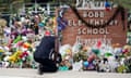 A Black man in a sleeveless T-shirt kneels with his head down in front of dozen of stuffed animals and flowers in front of a brick sign that says Robb Elementary School.