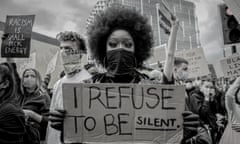 A young black protester with a big 'afro' hairstyle and a black bandana over her face holds a cardboard sign to the camera reading "I refuse to be silent"
