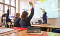 A teacher writes on a white board with three girls in a classroom with two raising their hands