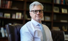 Middle-aged sort of smaller Latino man with coiffed gray hair, black-framed glasses, powder blue collared shirt, and light blue tie, sitting in leather chair in front of wall of bookcases.