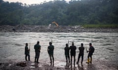 Seven men carrying spears stand by a river looking at a mechanical digger on the opposite bank