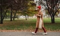 Mid-adult woman with mobile phone and cloth shopping bag walks in a park on an autumn day; leaves are seen on the ground. She wears a warm camel-coloured coat and knitted red hat, dark red trousers and white sneakers.