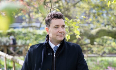 Edward Timpson stands in a park with new leaves and flowers, leaning on a wooden rail on the top of a wire fence.