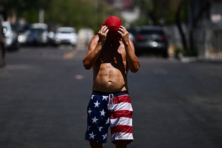 A man with no shirt and swim shorts in an American flag pattern, holds his head, in a ed ball cap, in the middle of a street with sun beating down on him.