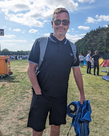 Smiling man, 50, with short brown hair and sunglasses; he wears a black Fred Perry top and black shorts plus a rucksack, and is holding a blue fold-up camping chair; people and tents can be seen in the park behind him