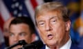 Older white man with orangey skin and poofy hair speaks into microphone in front of American flag.