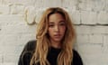 Tinashe, with long loose hair and wearing a black T-shirt standing against a white painted wall.