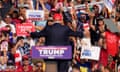 A man wearing a blue suit and red hat faces a crowd full of people wearing red, white and blue and holding signs in support of Donald Trump.