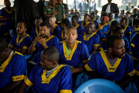 A young Black man with a shaved head looks directly at the camera, sitting among dozens of other, mostly older Black men and one white man, all wearing bright blue tunics with bright yellow stripes at the V-necks and the shirt sleeves. They sit in a shaded area in rows.