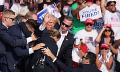 Trump waving to supporters with blood on his face, as security guards huddle around him, with crowds of supporters in the background