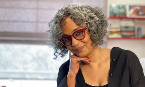 Arundhati Roy, with curly above-shoulder-level hair and wearing glasses and a button shirt over a T-shirt, rests her chin on her hand and smiles slightly