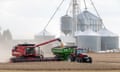 A red combine harvests soybeans and pours them into a green container on the back of a tractor, with grain silos in the background.