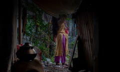 A middle-aged woman in a shalwar kameez [trousers and tunic] standing outside a corrugated iron-walled shack. A clay oven is in the gloomy interior