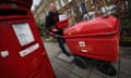 A Royal Mail delivery worker pushes a red mail cart down a street of Victorian houses in London, past a red postbox