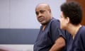 Bald and paunchy middle-aged Black man in navy blue prison top looks at camera, with painted cinder-brick walls beyond him.