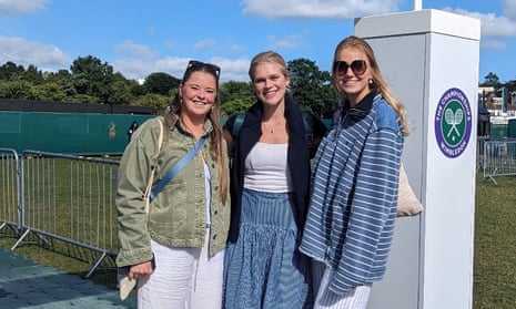 three young women, all with long fair or blond hair, standing at the entrance in front of metal fencing and a white pillar with the Wimbledon logo; one wears white with a khaki jacket, another a white top with a blue floaty skirt, and the third a blue and white striped sweatshirt over loose, striped white trousers plus large sunglasses. It is sunny and they are all smiling.
