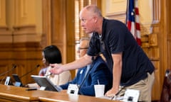 man wearing black shirt and khaki pants stands behind a table as two other people sit next to him