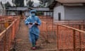 A health worker in a mask and surgical scrubs, holding small boxes of medicine, walks past plastic-betted fences and a corrugated iron roofed building in Africa