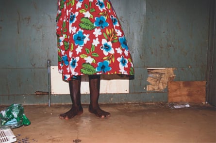 A view of Nancy Kidd from the waist down as she stands in front of a damaged wall in her house. She is barefoot and wearing a bright red skirt with blue and white flowers on it