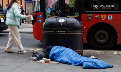 A homeless person sleeps on a pavement in a sleeping bag next to a bin, which a woman places litter in