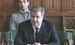 A middle-aged, grey-haired man in dark suit and tie sits on a green chair and speaks into a microphone