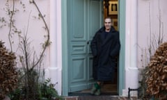 John Galliano in a big dark coat, hands in pockets, standing in the doorway of a house with a duck egg blue door and light pink facade