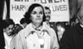 A black-and-white image of a white woman with coifed dark hair and a light knitted cap, and an overcoat with a large collar, chin up as she walks amid a crowd.