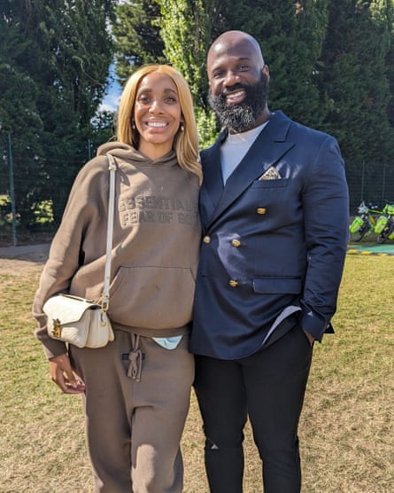 a smiling, happy-looking black couple in their 30s; she has long straight reddish hair and wears a loose, light brown tracksuit with hooded top; he has a shaven head and bushy beard and wears a smart navy double-breasted blazer with gold buttons over a white T-shirt and black jeans. They are standing shoulder-to-shoulder as if they have their arms around each other 