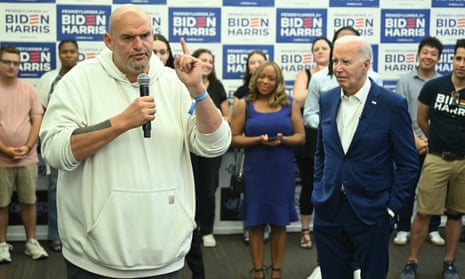 man wearing white hoodie holds microphones and gestures as people including man wearing blue suit look at him