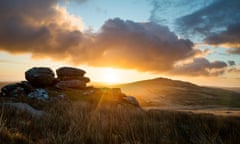 The sun shines through clouds over a distant hill with boulders in the foreground