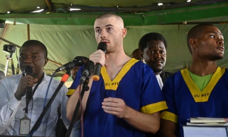 A young white man with his head shaved, wearing a bright blue shirt with a broad yellow stripe at the V-neck and shirtsleeves, stands among a group of other people, all Black, speaking into a microphone and gesturing with one hand.