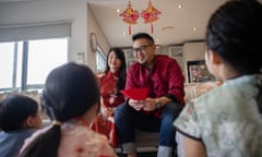 A mother and father sitting on a couch holding red packets of money for lunar new year; their three children sit before them on the floor