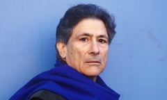 A man in a blue scarf against a blue background looks  into the camera.