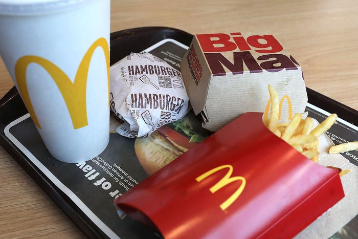 A Big Mac, a hamburger, a fountain drink, and french fries sit on a black plastic tray while packaged in McDonald's-branded wrappers.