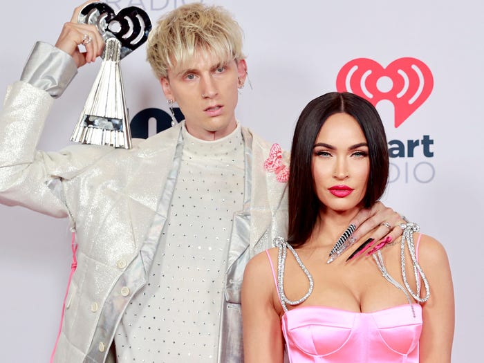 Rapper Machine Gun Kelly wears a white and silver suit and holds an iHeart Radio Music award while posing with his hand on girlfriend Megan Fox's neck, who is wearing a pink satin jumpsuit and staring at the camera.