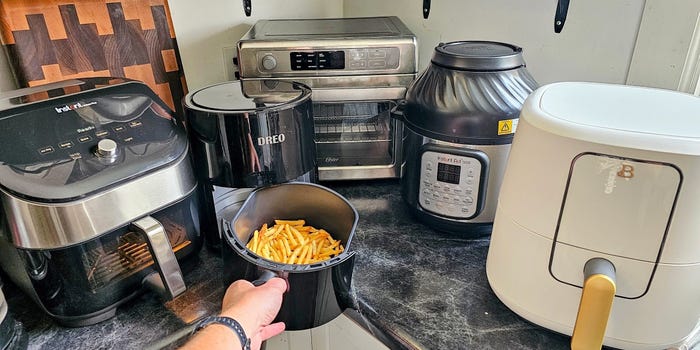 Five air fryers are lined up on a counter while a hand holds onto an air fryer basket full of french fries.