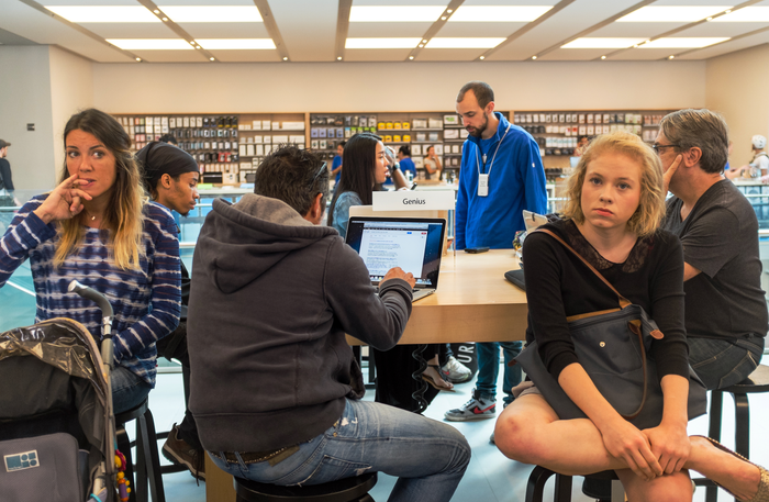 Apple customers sit on stools around a wooden table, waiting for their Genius Bar appointments.