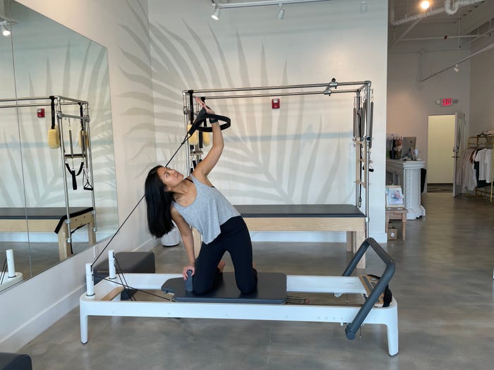 The writer kneels on a Pilates machine carriage and extends an arm holding a rope connected to the machine above her head